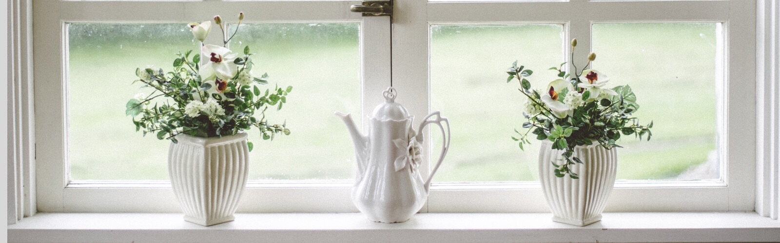 Two white vases filled with flowers and a white teapot aligned on a windowsill.